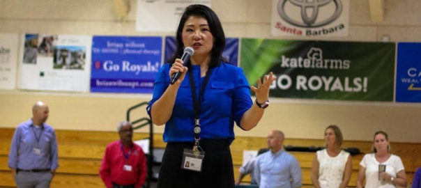 Woman in blue shirt holds a microphone and talks in a gym