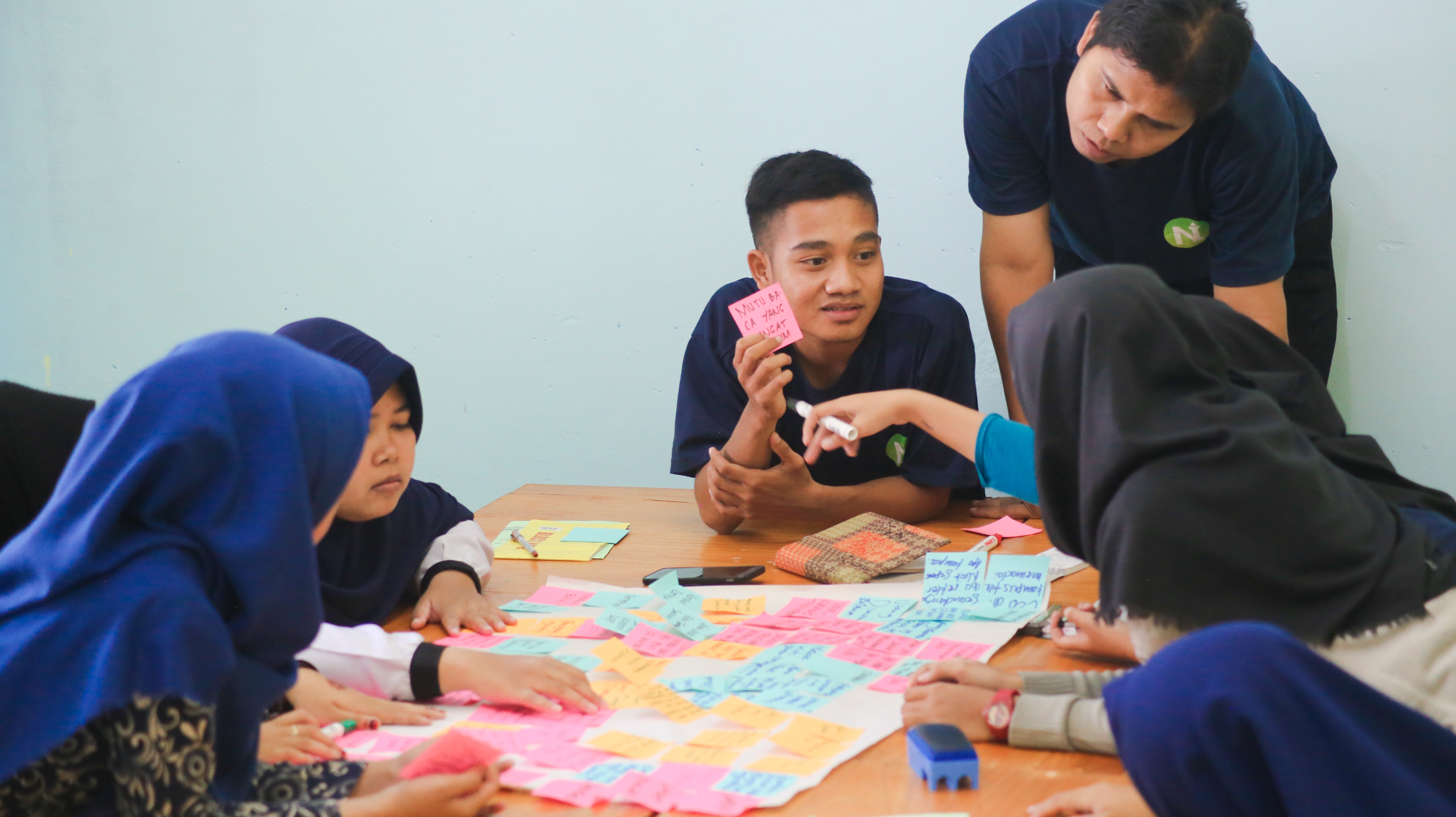 Seven people sit around a table looking and holding up colorful post-it notes. Several women wear a headscarf, two blue and two black.