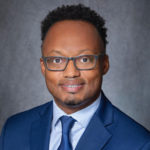 Profile picture black man with glasses wearing a dark blue suit and dark blue tie.