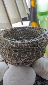 A small completed pine needle basket used for storing small knick-knacks perched on white seashells.