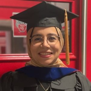 Woman wearing hijab, glasses, and black graduation cap and gown..